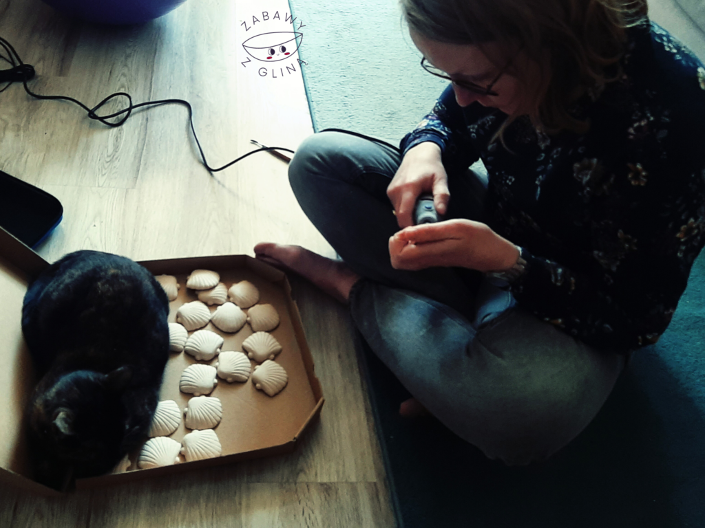 Kobieta elektrycznym narzędziem szlifuje figurki ceramiczne muszelek. Obok w pudełku na pizzę leżą kolejne muszelki ceramiczne. Na muszelkach siedzi ciemny kot.